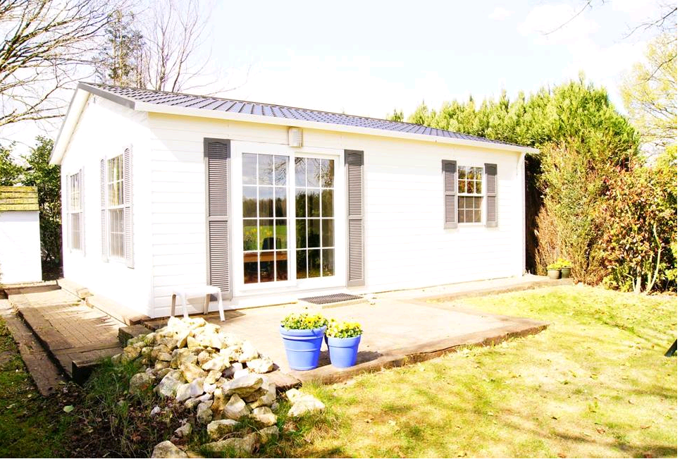 Een wit houten chalet met een terras en een grasveld, omringd door beplanting.