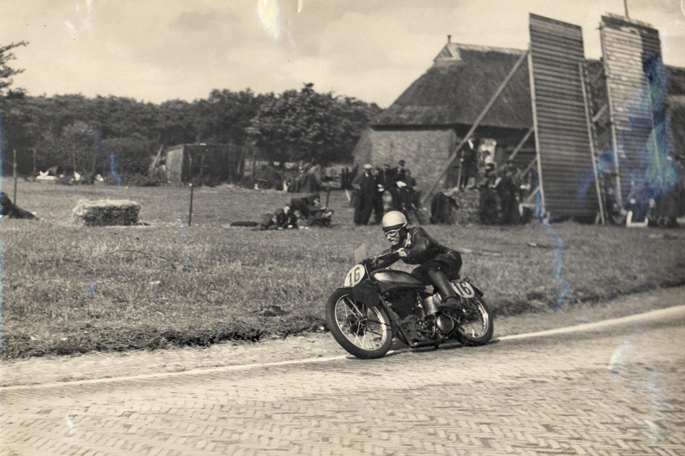 Historische foto van een motorrijder die tijdens één van de eerste TT's door een dorp racet.