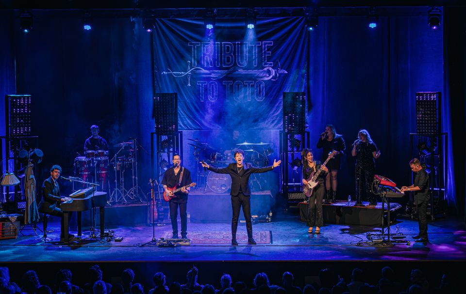 Foto van de band met hun instrumenten op een podium in paars-blauw licht.