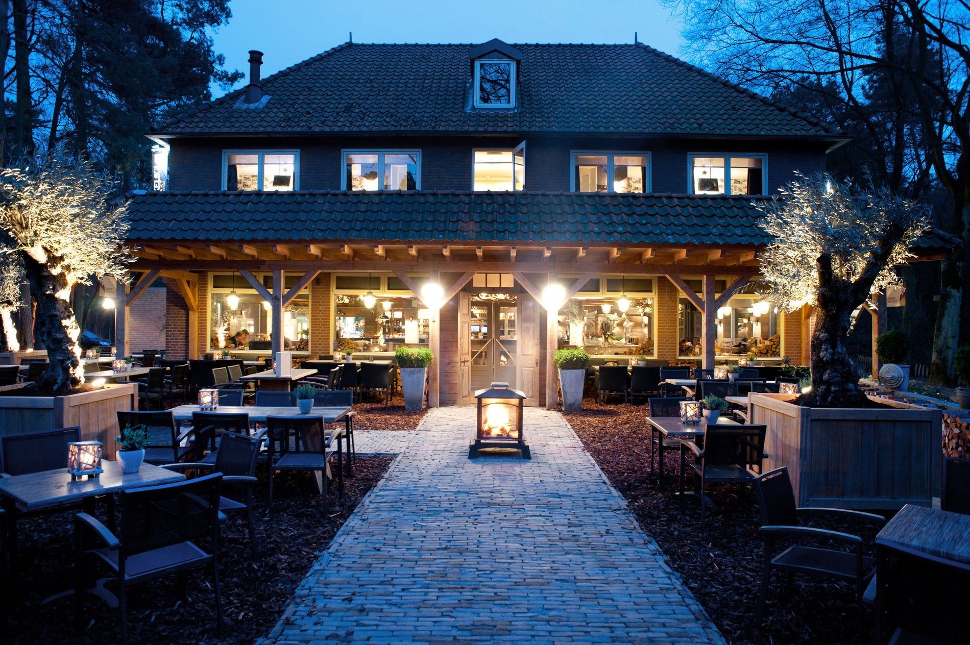 Terras in bosrijke omgeving met het hotel-restaurant op de achtergrond in de avond met verlichting.