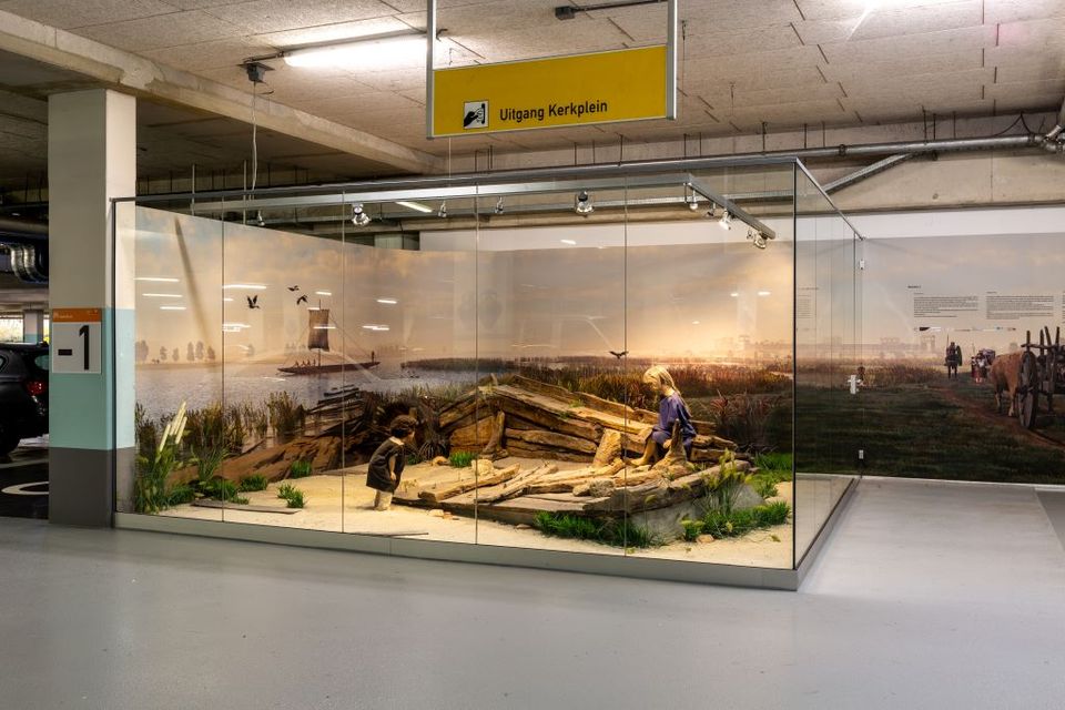 Vitrine in de parkeergarage met daarin de resten van een schip en twee poppen die kinderen verbeelden.