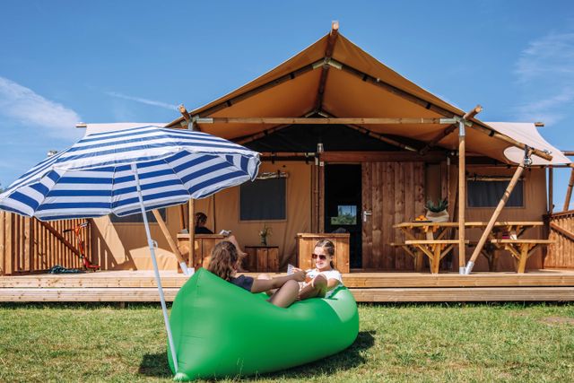 Kinderen ontspannen voor een ruime glampingtent op Vakantiepark De Heigraaf. De safaritent heeft een houten veranda en biedt comfortabel kamperen in de buitenlucht.