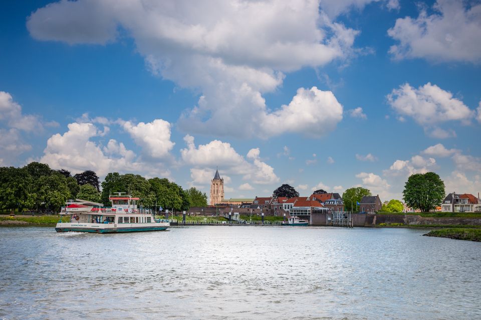 Een waterpartij met daarop een veerpont, op de achtergrond een vestingstad met daar bovenuit een hoge kerktoren.