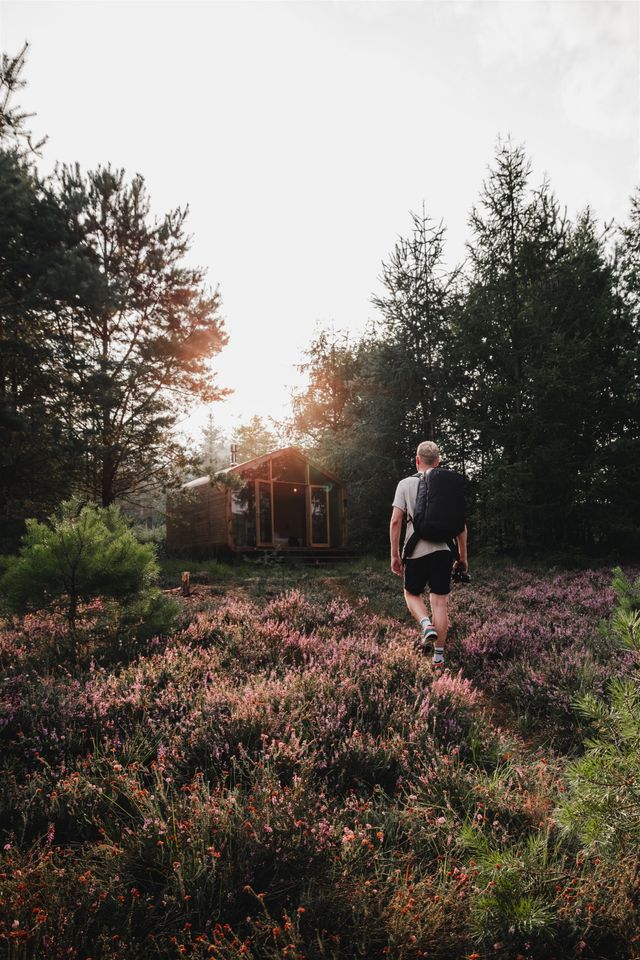 Een man loopt met zijn backpack op door paarse heide richting een cabin.