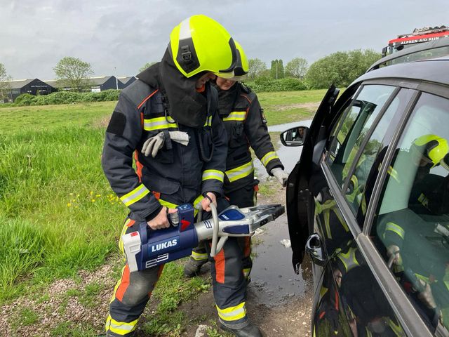 Twee brandweermannen waarvan een gereedschap vast houd en ze staan naast een auto.