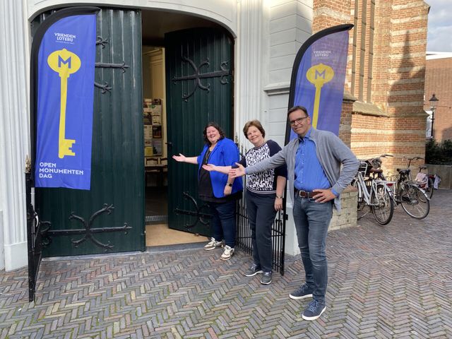 Drie vrijwilligers verwelkomen bezoekers bij de ingang van een monumentale kerk tijdens Open Monumentendag in Woerden, met vlaggen aan weerszijden van de deur.