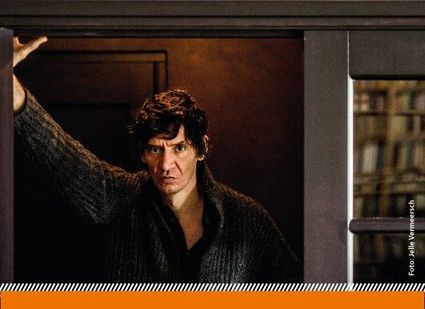 Donker getinte filmstill van een man met intense blik die in een deuropening staat en één arm omhoog houdt tegen het kozijn. Op de achtergrond zijn vage boekenkasten zichtbaar, wat een gespannen en introspectieve sfeer oproept.