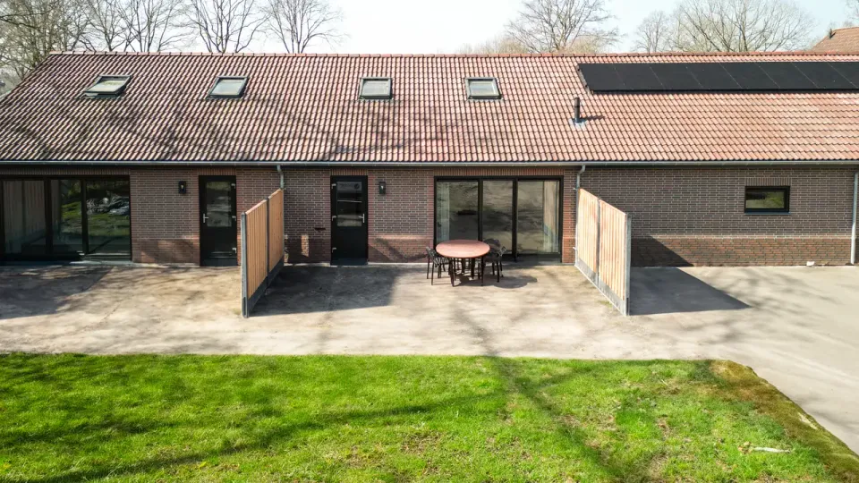 Zicht op een voormalig boerenschuur, bedekt met rode dakpannen en diverse dakraampjes. De schuur is d.m.v. houten schotten opgedeeld in twee B&B units. Groen grasveld voor de units en terras.