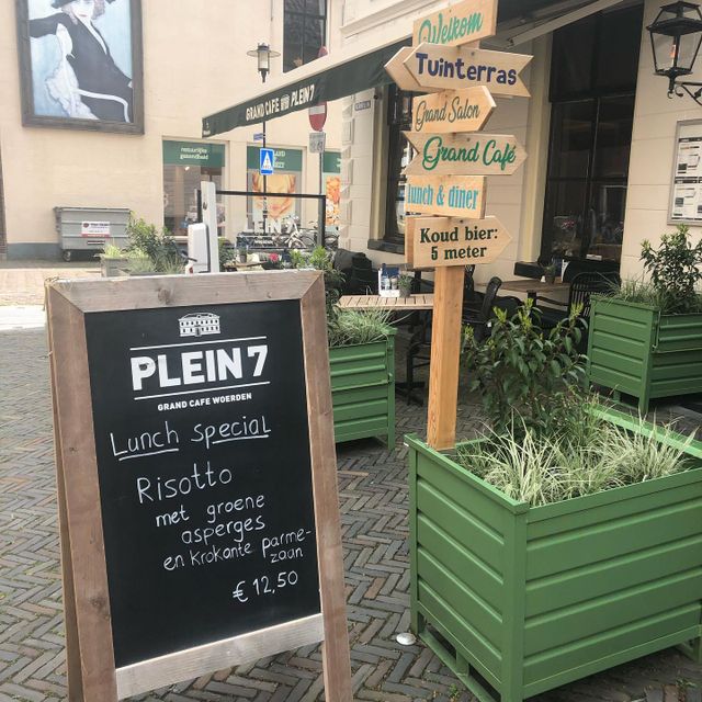 Een uitnodigend terrasbeeld van Grand Café Plein 7 in Woerden, waar een handgeschreven krijtbord de lunchspecial van de dag aankondigt. Met groene plantenbakken, sfeervolle wegwijzers en een zonnige setting straalt de plek warmte en gastvrijheid uit — perfect voor een smakelijke tussenstop in het hart van de stad.