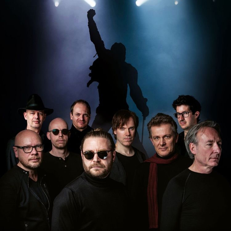 Donkere foto van de uit 9 mannen bestaande band in donkere kleding met op de achtergrond een silouet van een persoon die een optreden geeft.