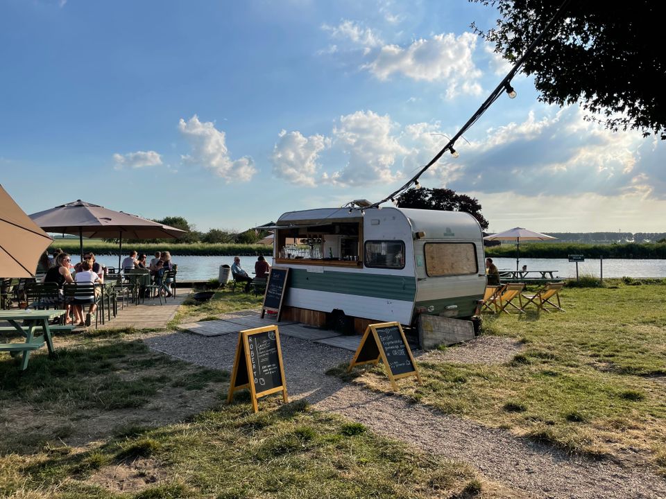 Terrasje aan de Vecht bij Buitenplaats op 't Eiland in Nederhorst den Berg. Vanuit de caravan worden drankjes geserveerd en mensen zitten op strandstoelen en zitjes aan de Vecht.