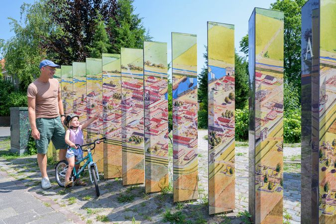 Een vader en dochtertje op een loopfietsje kijken naar het kleurrijke kunstwerk dat Forum Hadriani herdenkt.