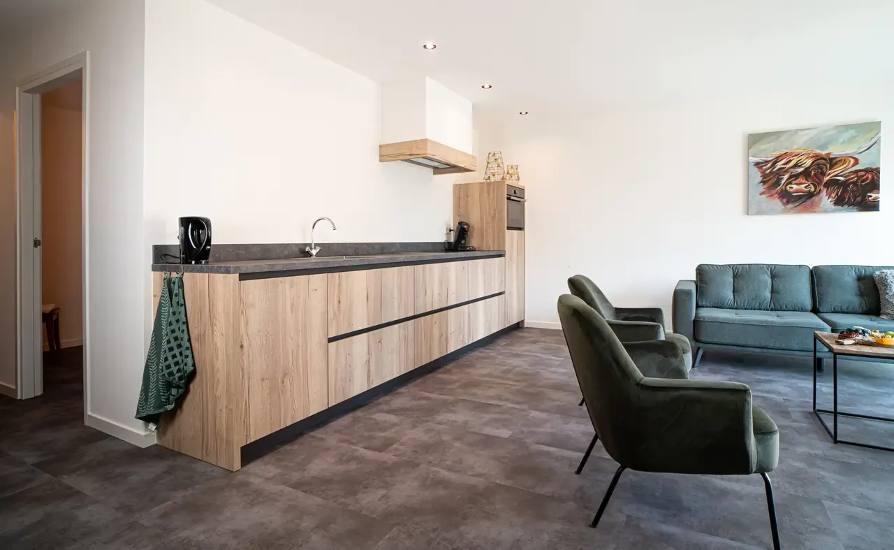 Zicht op een houten keukenblok en zithoek met lichtgrijze bank en donkergroene kuipstoeltjes, Boven de bank hangt een schilderij met een koeienkop.