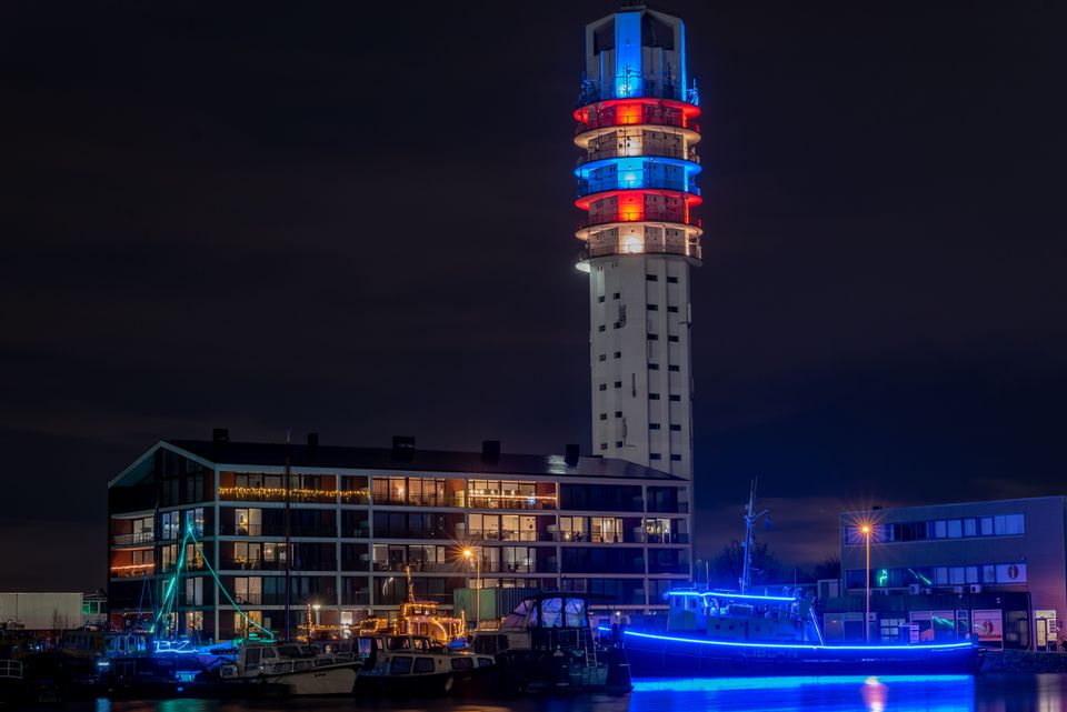 de lichttoren schijnt de kleuren blauw, rood en groen met op de voorgrond ook nog 2 boten die verlicht zijn door een lichtslinger lans de contouren van de boot.