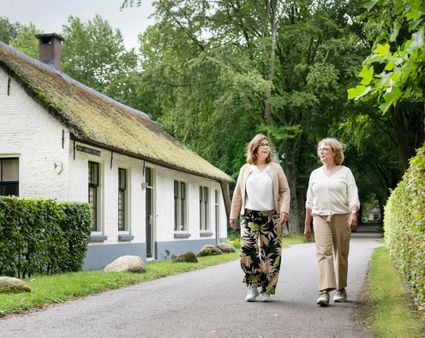 Twee dames met aan de linkerkant het witte ambtenarenhuisje in Frederiksoord. Het ambtenarenhuisje is een monument.