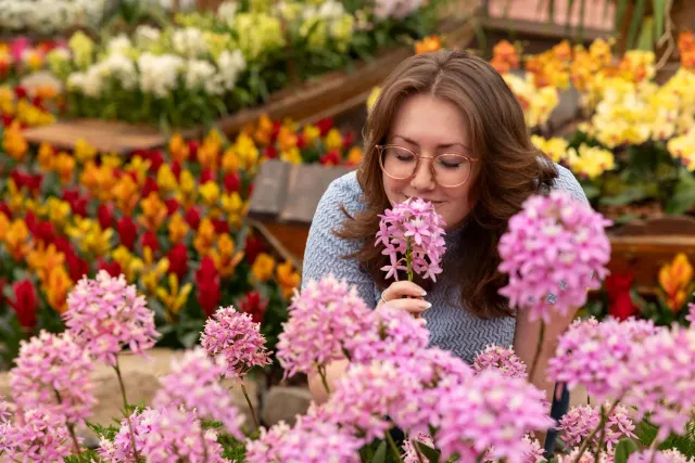 Vrouw staat tussen allemaal bloemen en ruikt aan een mooie roze bloem