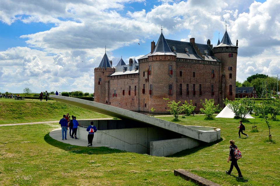 Buitenaanzicht van kasteel Muiderslot met op de voorgrond het Waterschild en een aantal bezoekers die in de kasteeltuin lopen.