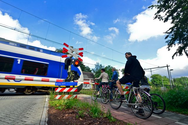 Fietsers wachten bij een gesloten spoorwegovergang terwijl een passagierstrein voorbijrijdt. De foto laat de combinatie van fietsen en openbaar vervoer in de regio zien.
