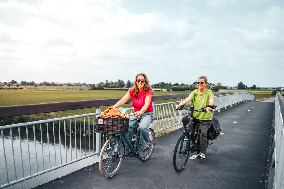 Fietsbrug in de Utrechtse Waarden bij IJsselstein, Groene Hart. Twee fietsers rijden over een moderne brug boven het water, omringd door uitgestrekte polders, weilanden en een weids landschap onder een bewolkte lucht.