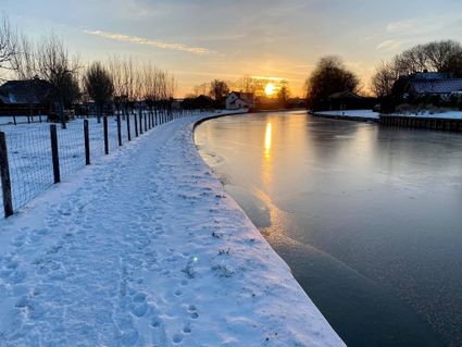 Een verstilde winterochtend langs de Oude Rijn, waar een besneeuwd wandelpad en een dun laagje ijs op het water samenkomen in het zachte licht van de opkomende zon. De warme gloed weerspiegelt prachtig op het bevroren oppervlak en geeft het winterse landschap een serene, bijna magische sfeer.