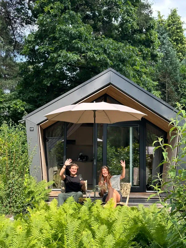 Een stel zit op hun terras voor hun houten huisje, een Diamondsuite, op vakantiepark KampinaStaete in Oisterwijk.