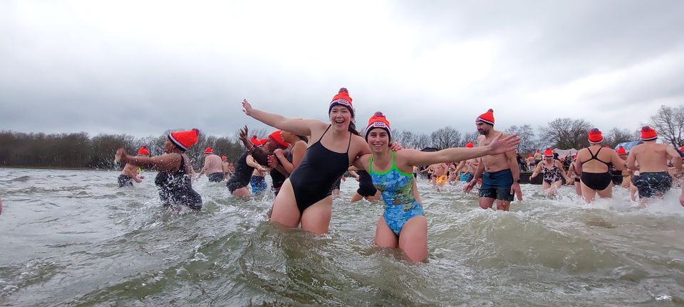 De afbeelding toont deelnemers aan de nieuwjaarsduik. Op de voorgrond staan twee vrouwen met gespreide armen in het water. Op de achtergrond zijn meerdere deelnemers, winterse lucht en logo’s van Reddingsbrigade Oss.