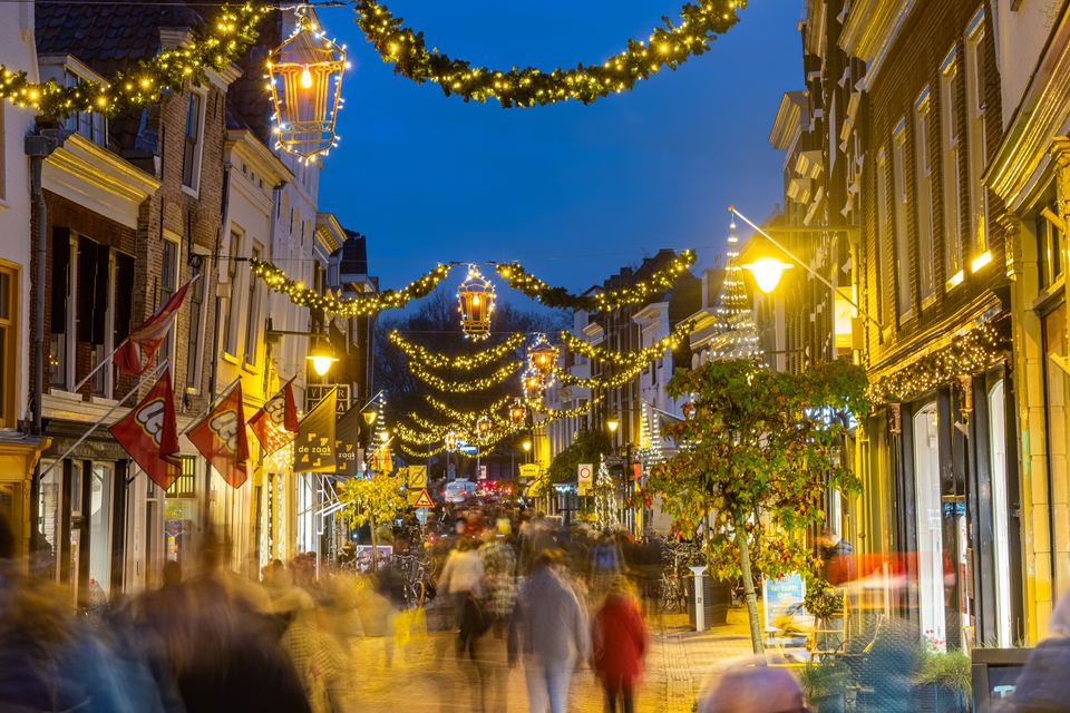 Een drukke winteravond in de stad: onder fonkelende lichtslingers vullen winkelende mensen de straat, terwijl historische gevels baden in warm licht. De beweging van het publiek en de feestelijke verlichting geven het beeld een levendige, bijna magische sfeer waarin winterse gezelligheid en stadse dynamiek samenkomen.