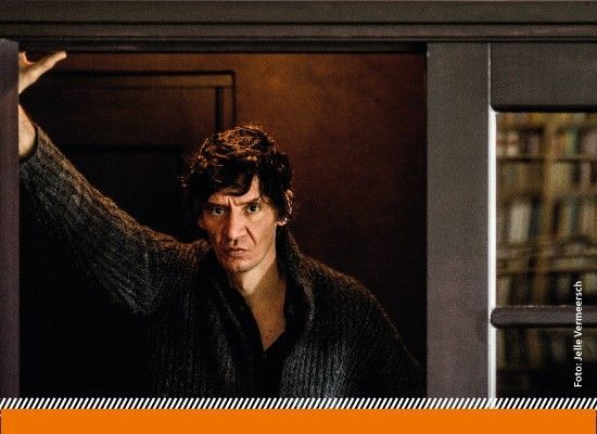 Donker getinte filmstill van een man met intense blik die in een deuropening staat en één arm omhoog houdt tegen het kozijn. Op de achtergrond zijn vage boekenkasten zichtbaar, wat een gespannen en introspectieve sfeer oproept.