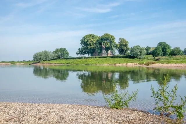 Zicht op een plas met groene oevers, een huis tussen de bomen op de achtergrond.