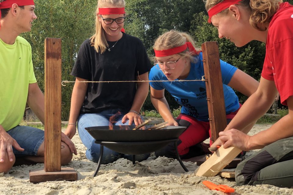 Vier jongeren zitten rondom een vuurschaal en proberen het vuur aan te wakkeren.