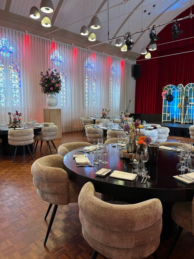 Ronde tafels met donker tafelblad zijn gedekt met glazen, servetten en bestek; in het midden staan bloemstukken. De zaal heeft hoge ramen met gekleurd glas en sfeerverlichting.