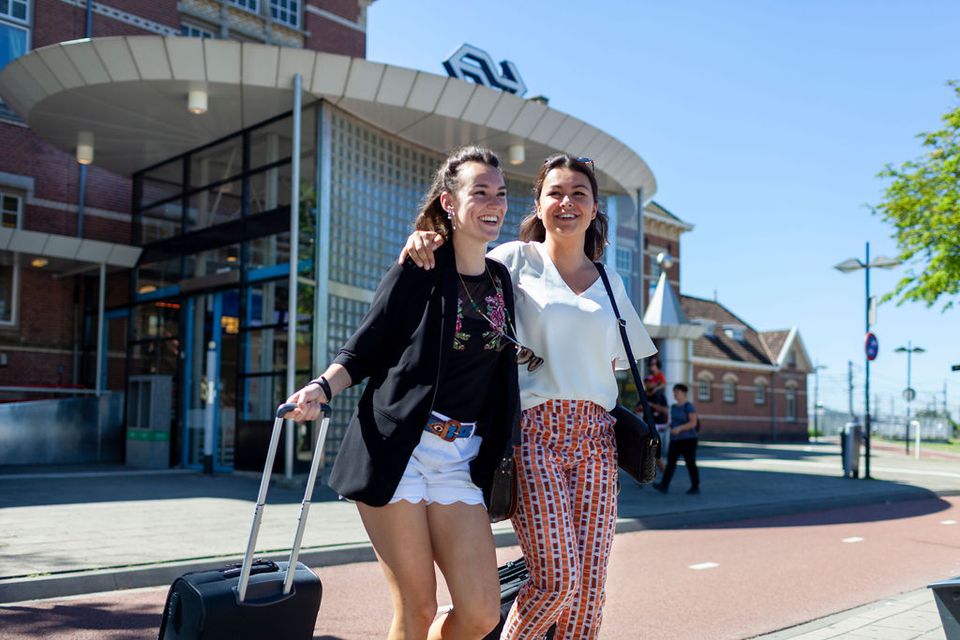 Treinstation Woerden, Groene Hart. Twee reizigers met rolkoffers lopen voor het stationsgebouw en starten hun bezoek aan de historische vestingstad, goed bereikbaar met trein en openbaar vervoer.
