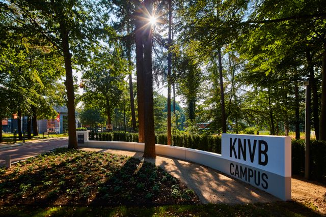 Entree van de KNVB Campus in Zeist tussen de bomen, met het herkenbare KNVB Campus-bord. De foto geeft een sfeervolle indruk van de groene ontvangst en toegang tot het sportcomplex.