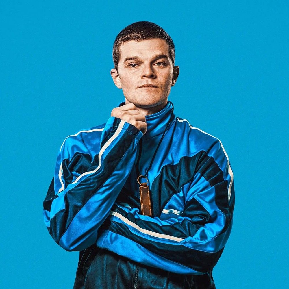 Foto van een jonge man in een blauw gestreept trainingsjack tegen een lichtblauwe achtergrond.