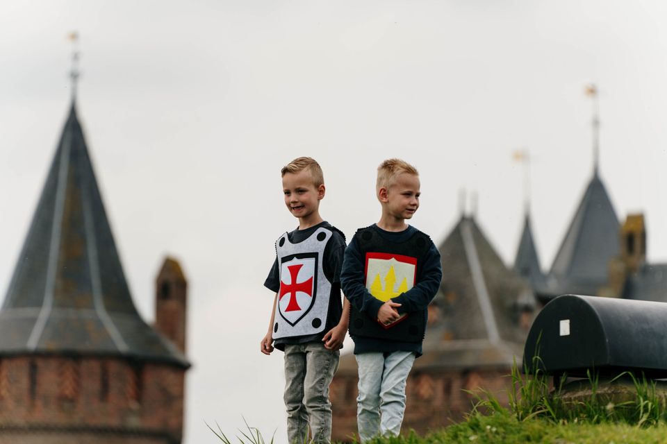 Twee jonge kinderen met ridderkostuum staan op een heuvel in de kasteeltuin met op de achtergrond vaag zichtbaar Kasteel Muiderslot in Muiden.