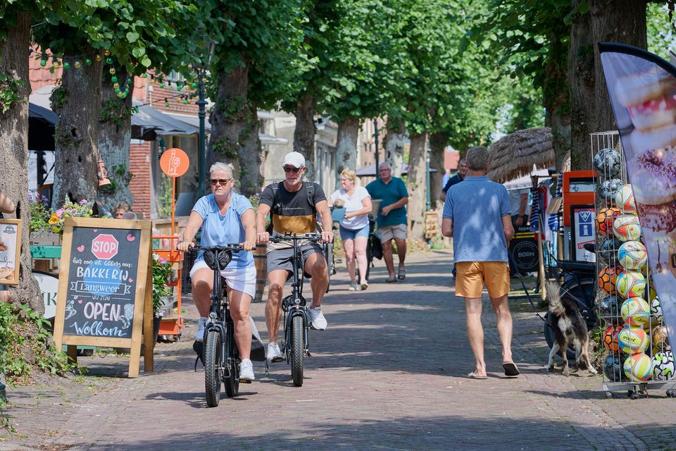 Een oudere man en vrouw op elektrische vouwfietsen door de winkelstraat van Langweer. Op de achtergrond twee lopende fietsers en een man met een hond. Aan de zijkanten staat een bord van de bakker en voetballen in een rek.