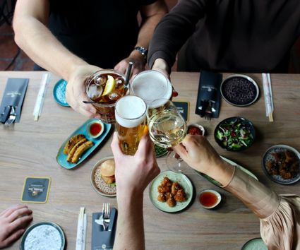 Aan een tafel zitten twee mensen, zij proosten met een drankje en de tafel is gedekt met hapjes.