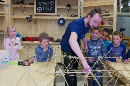 Begeleider van Ontdekstation laat aan kinderen zien hoe je met houtjes een constructie bouwt.