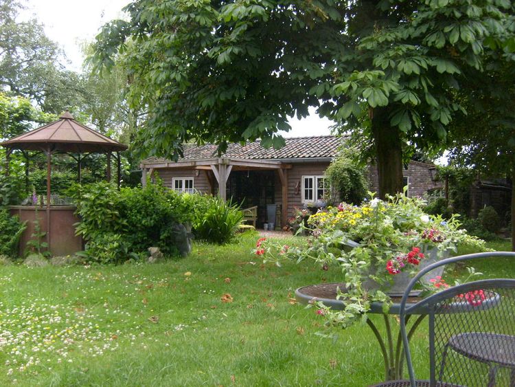 Zicht op een houten chalet met afdak op een groen grasveld met aan de linkerkant een 6-hoekige volière en op de voorgrond rechts een smeedijzeren zitje met kleurrijke bloemen.