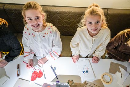 Twee jonge meisjes zitten aan een tafel en laten hun zelfgemaakte tekeningen zien, omringd door stiften, papier en knutselmateriaal.