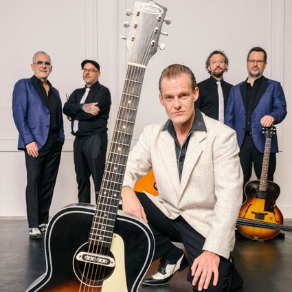 Foto van de band, 5 heren met hun muziekinstrumenten in blauw-zwart-wit gekleed, voor een lichte achtergrond.