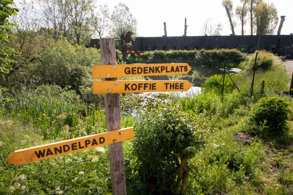 Een wegwijzer met gele bordjes eraan die verschillende richtingen op wijzen. Op één staat gedenkplaats, op die daaronder koffie thee en de onderste benoemt een wandelpad. Op de achtergrond zie je veel groen en een betonnen fort.