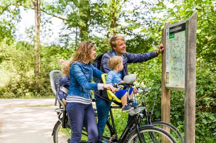Man en vrouw staan samen met de kinderen die op dfiets zitten te kijken naar een fietsroutebord.