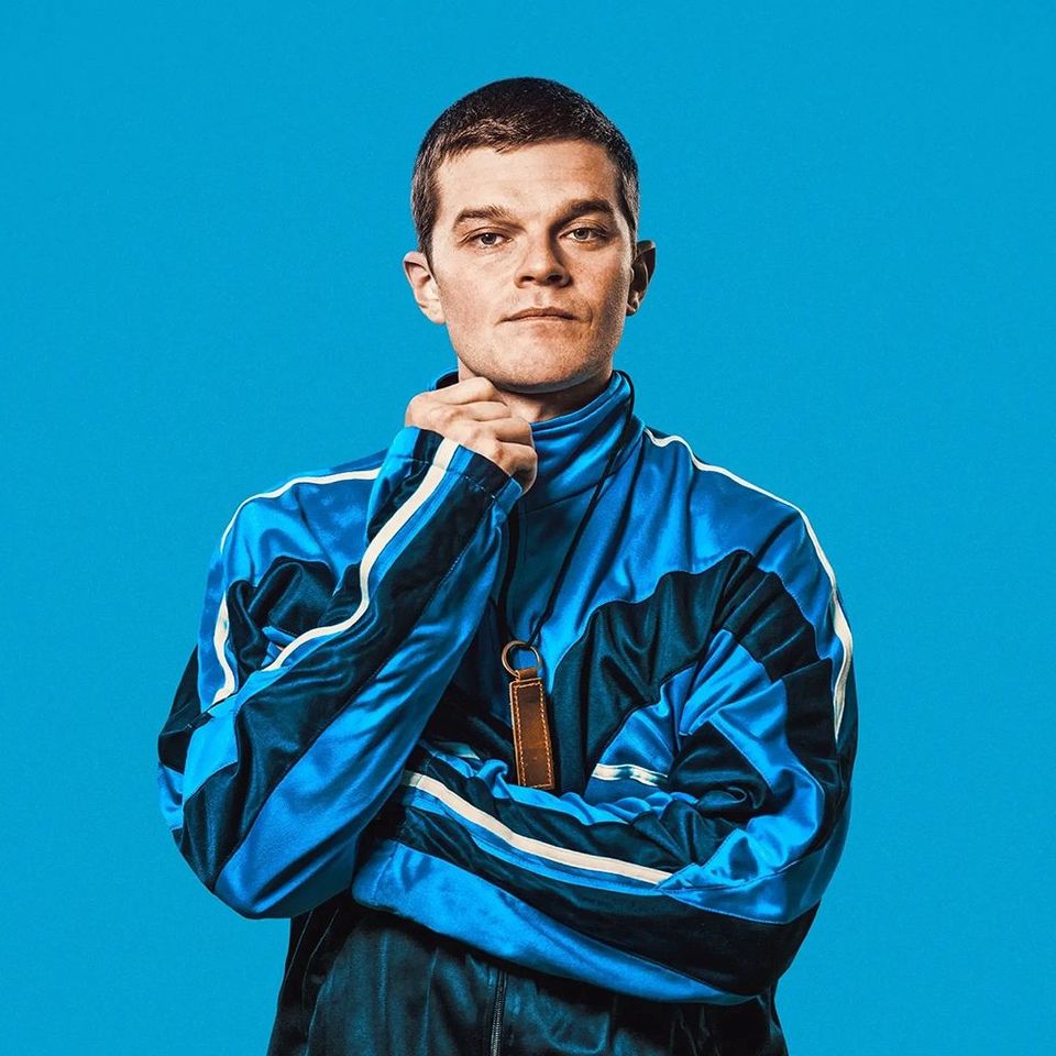 Foto van een jonge man in een blauw gestreept trainingsjack tegen een lichtblauwe achtergrond.