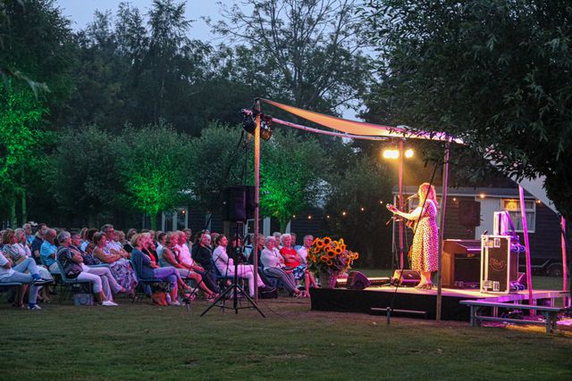 Publiek geniet van een live optreden tijdens het Midzomer Theater op Buitenplaats Kameryck, in een sfeervolle buitenomgeving met lichtdecor en bomen.
