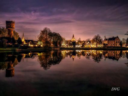 Een betoverend avondbeeld van Woerden, waarin de verlichte watertoren, kerktorens en historische gebouwen zich haarscherp spiegelen in het stille water van de singel. De paarse gloed van de lucht en de warme stadslichten creëren een bijna sprookjesachtige sfeer, waarin de stad haar mooiste licht toont.