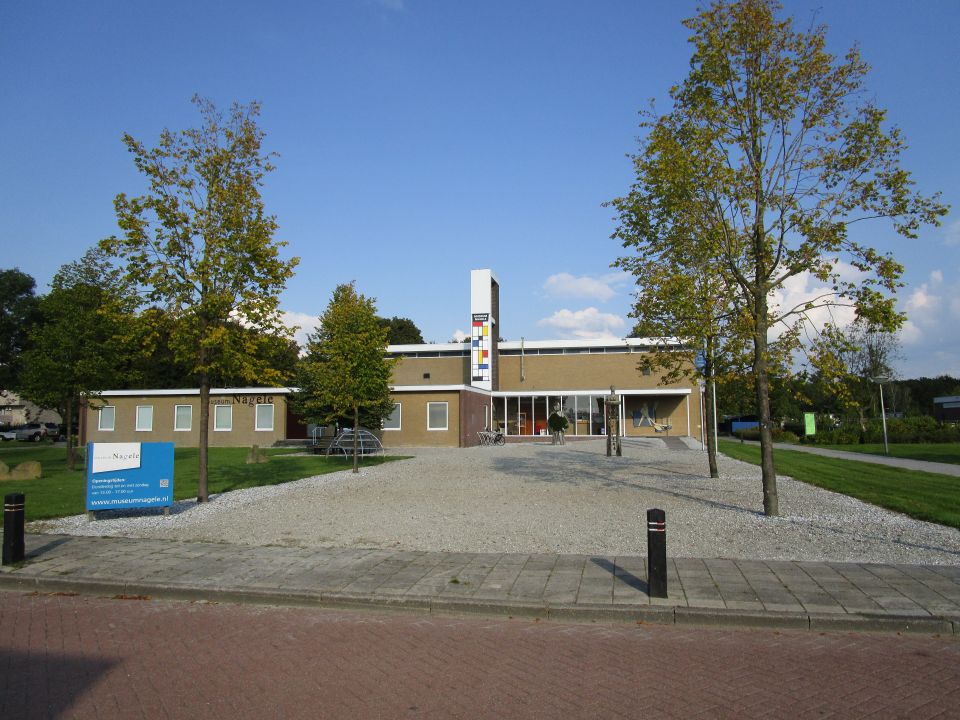 Het vooraanzicht van een gebouw met een plat dak. Om het gebouw heen staan diverse bomen. In Nagele in de Noordoostpolder.