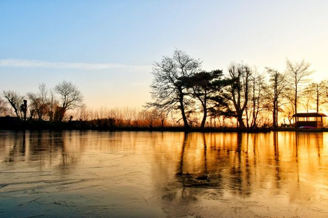 Sfeerbeeld van een bevroren plas bij zonsondergang in Bodegraven-Reeuwijk, met silhouetten van kale bomen en een theehuisje aan de oever in het Groene Hart.