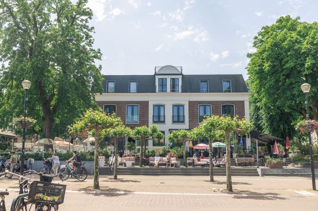 Oud gebouw in het centrum van Soest op de Utrechtse Heuvelrug. Er is een terras, fietsende mensen, groene bomen en het is zonnig.