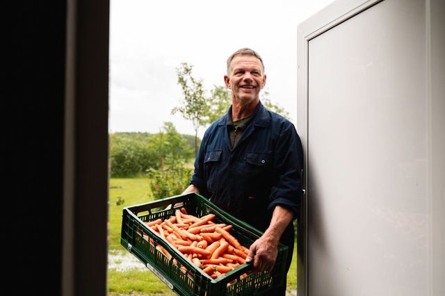 Ein Mann in Latzhose steht mit einer Kiste Karotten im Türrahmen.